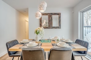 Uma sala de jantar com uma mesa de madeira posta para seis pessoas, rodeada por cadeiras, com luminárias pendentes geométricas no teto e um espelho decorativo acima.