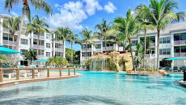 Une piscine d’un complexe hôtelier, avec une eau cristalline, entourée de palmiers et bordée d’un bâtiment blanc doté de balcons.