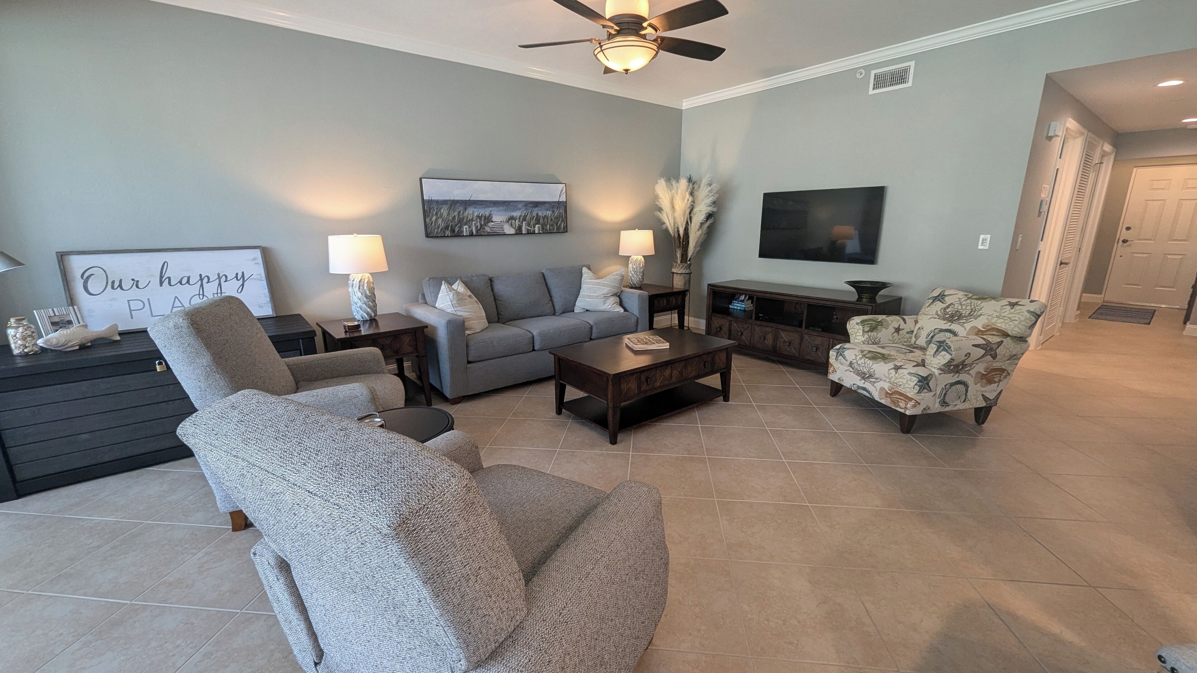 A living room with a grey sofa, a patterned armchair, a coffee table, and a TV on the wall.