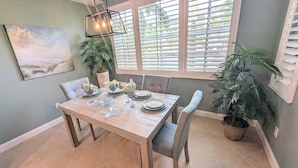 Un coin repas avec une table en bois dressée pour quatre personnes, entourée de chaises rembourrées. On y trouve une grande fenêtre avec des stores, une plante en pot et un tableau accroché au mur.