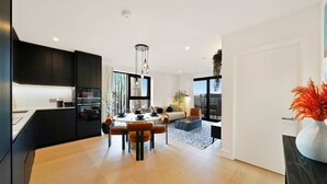 A modern kitchen with dark cabinetry, a dining area with a round table and orange chairs, and a living room with a sofa and a view of the city.