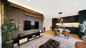A modern living room with a flat-screen TV, a dining area with a round table and chairs, and a kitchen area with a dark countertop and hanging lights.