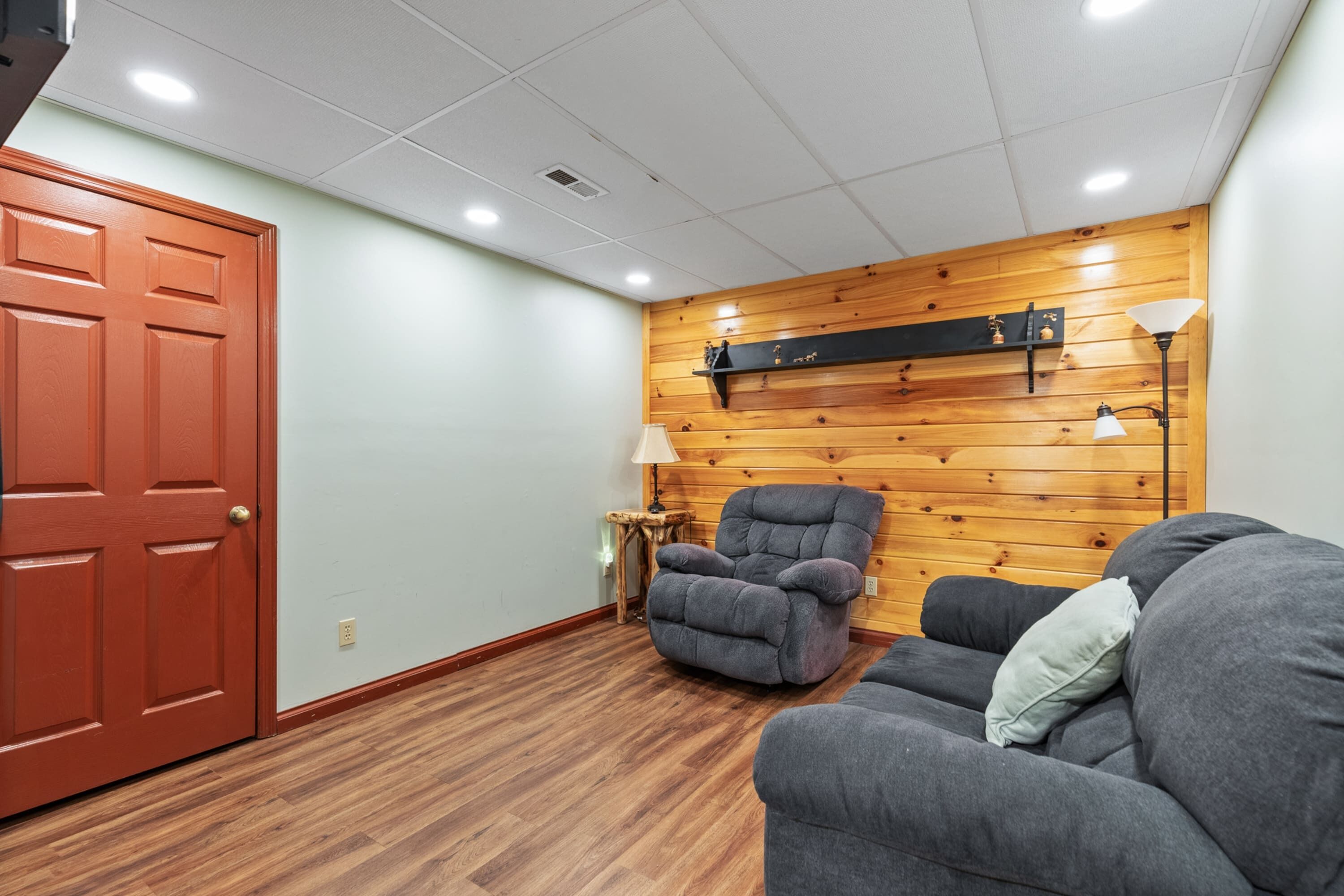 A room with a wooden wall, a grey sofa, a red door, and a lamp.