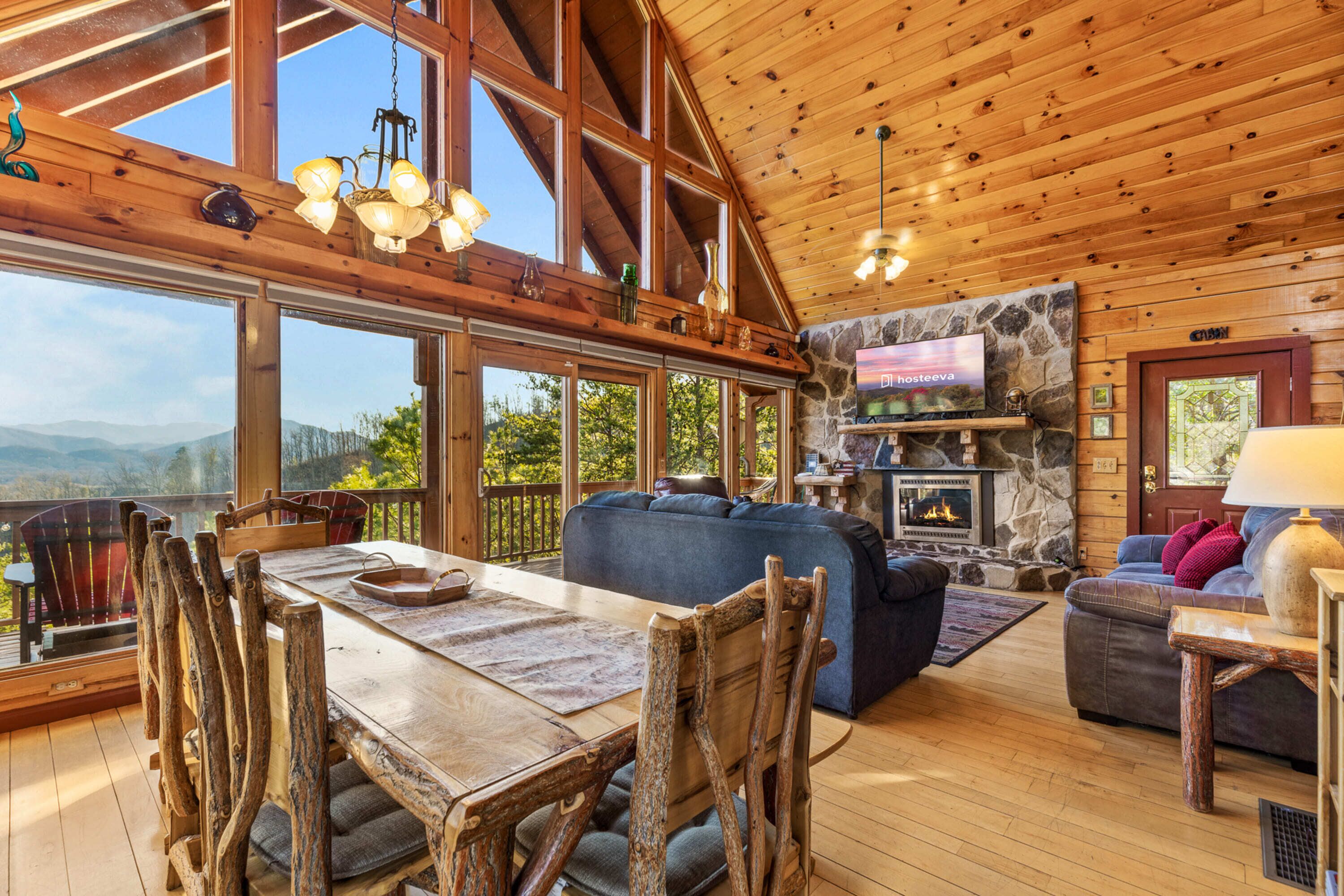 A spacious living room with a large wooden dining table, rustic chairs, a stone fireplace, and a stone wall TV.