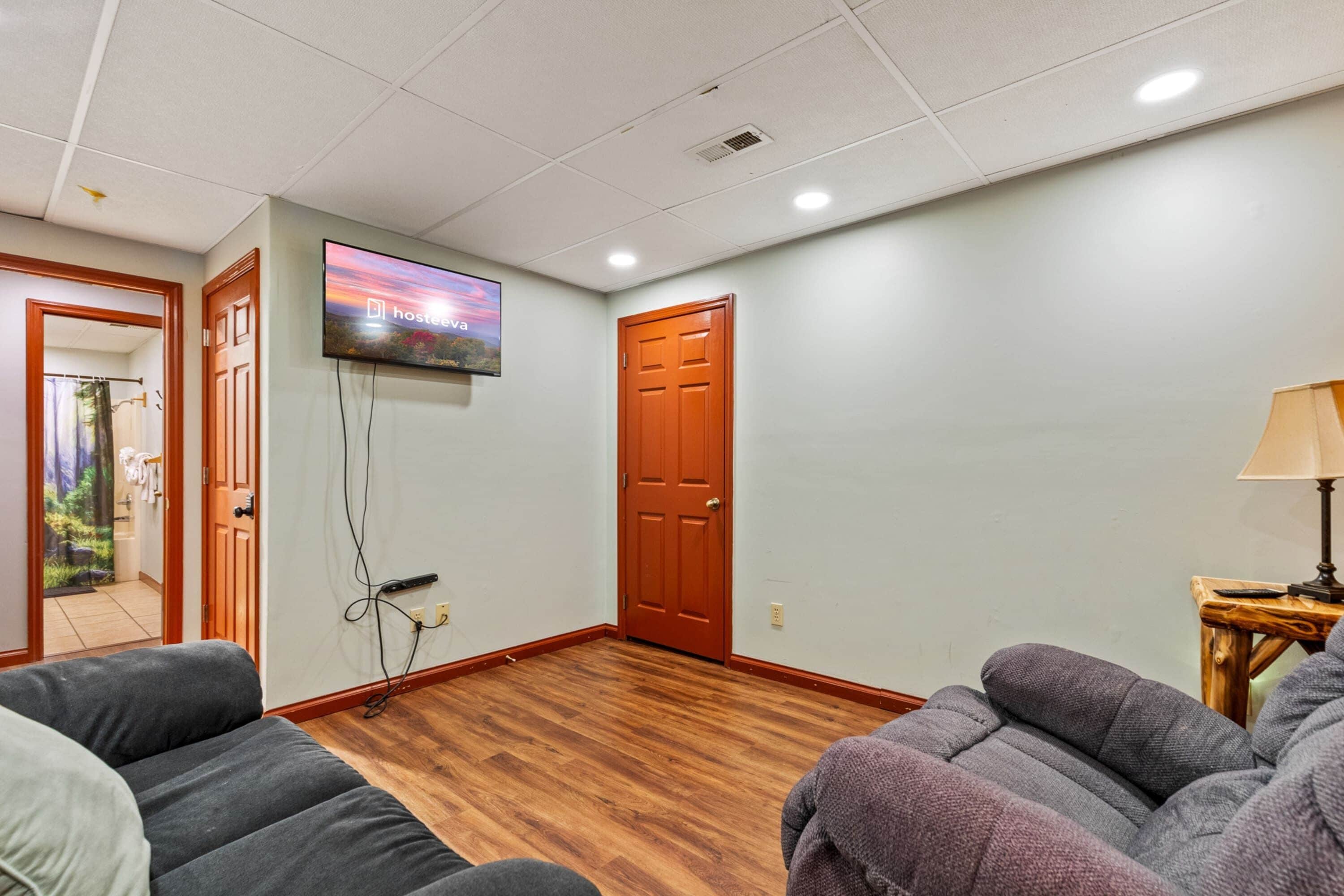A hotel room with a wooden floor, a television mounted on the wall, a door, and two sofas.
