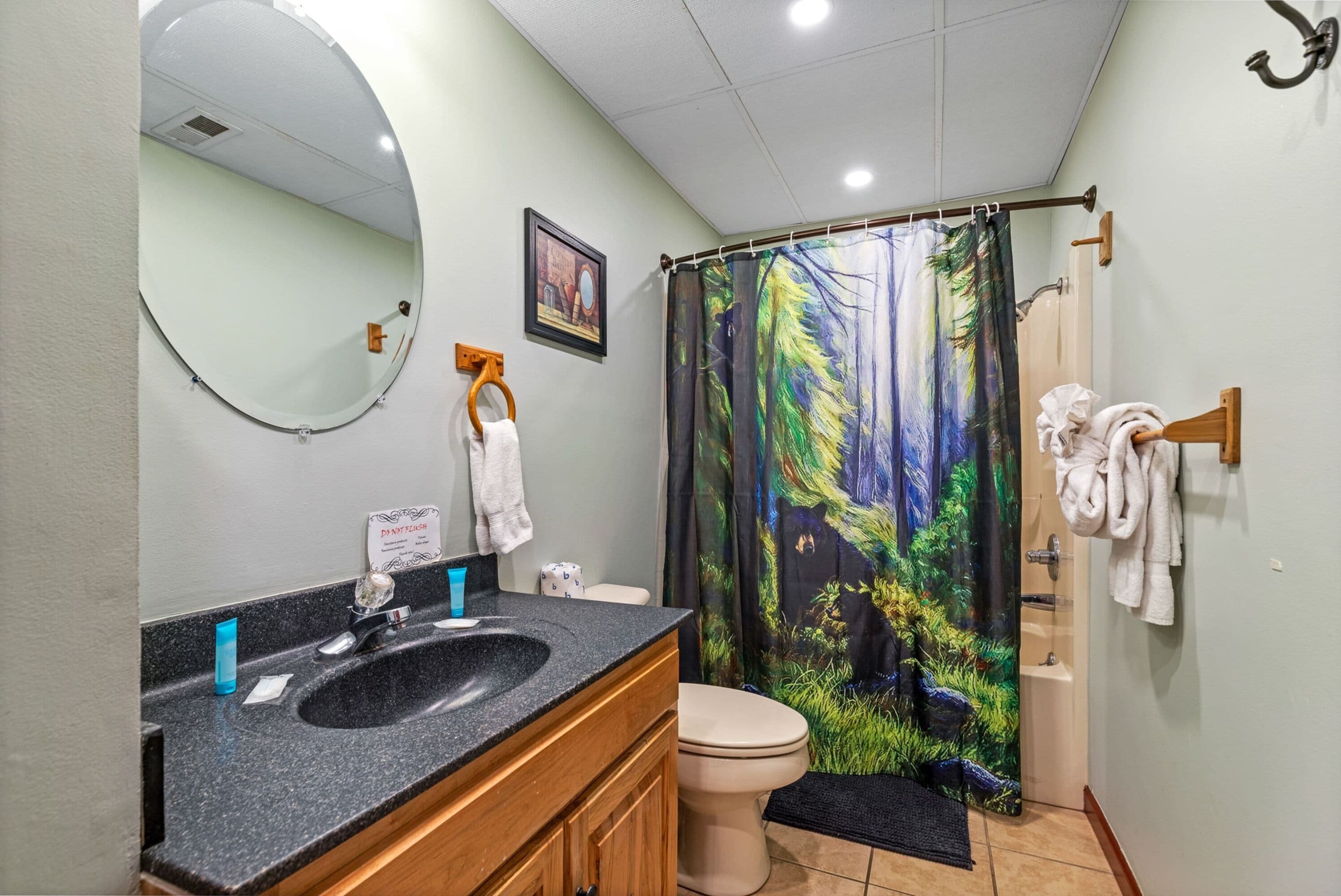 Une salle de bain avec un miroir rond, un lavabo avec un meuble en bois, des toilettes et un rideau de douche orné d’un motif représentant une forêt.