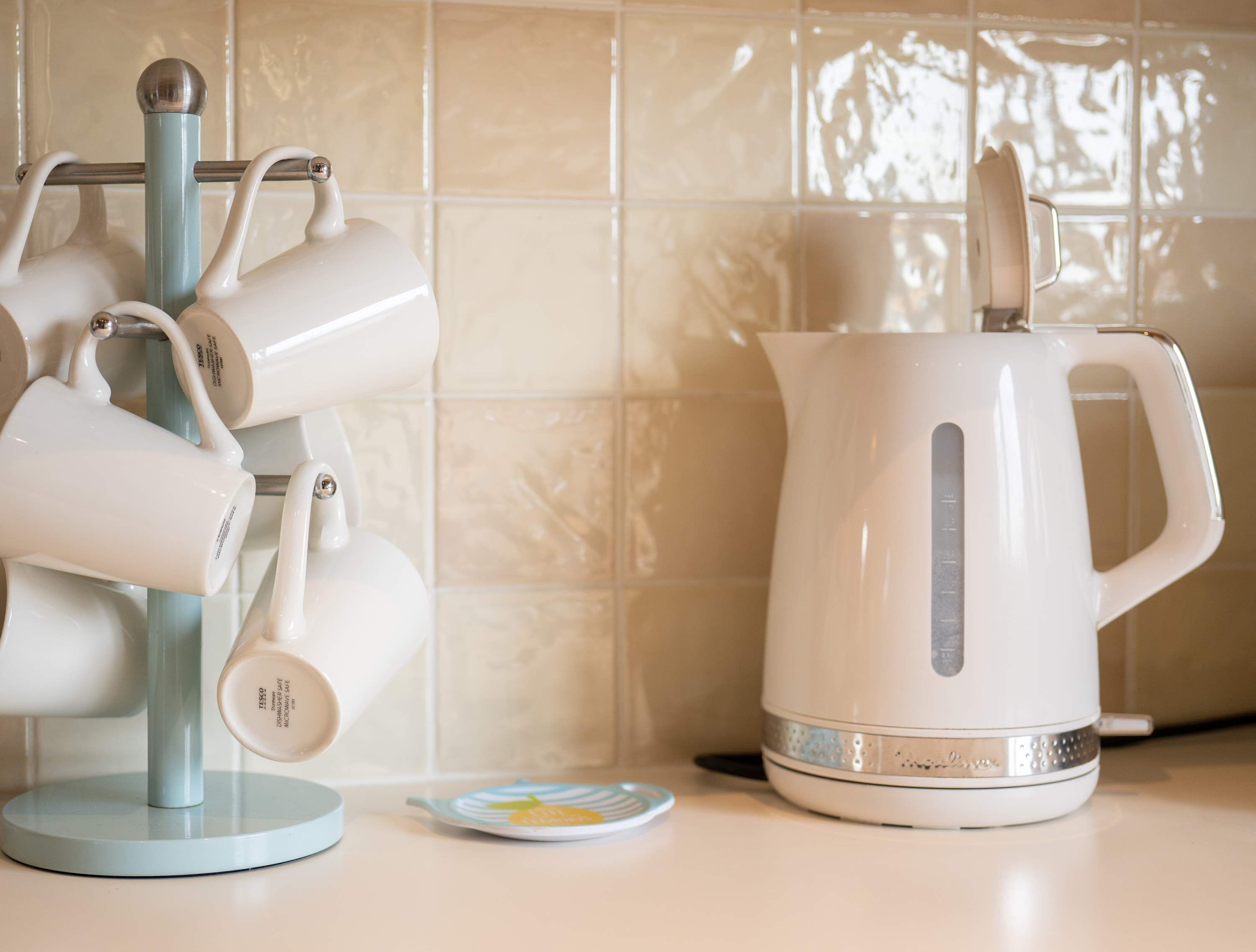 Una cocina con un hervidor blanco y un soporte para tazas blancas frente a un rebozado de azulejos.