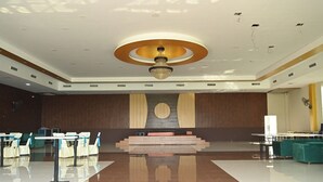 A spacious conference room with a central stage, tiered seating, and a chandelier. - NRI Hotel (Barara)