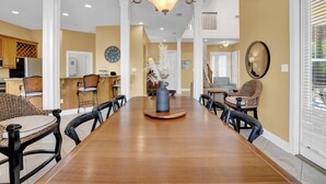 A spacious dining area with a long wooden table, multiple chairs, a chandelier, and a view of the kitchen and living area in the background.