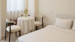 Een slaapkamer met een groot bed, een kroonluchter, een eettafel met stoelen en een raam met dunne gordijnen.
