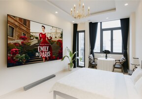 Habitación de hotel con un cartel grande de 'Emily en París', una cama, una mesita con sillas, una planta en maceta y vistas de la ciudad a través de amplios ventanales.