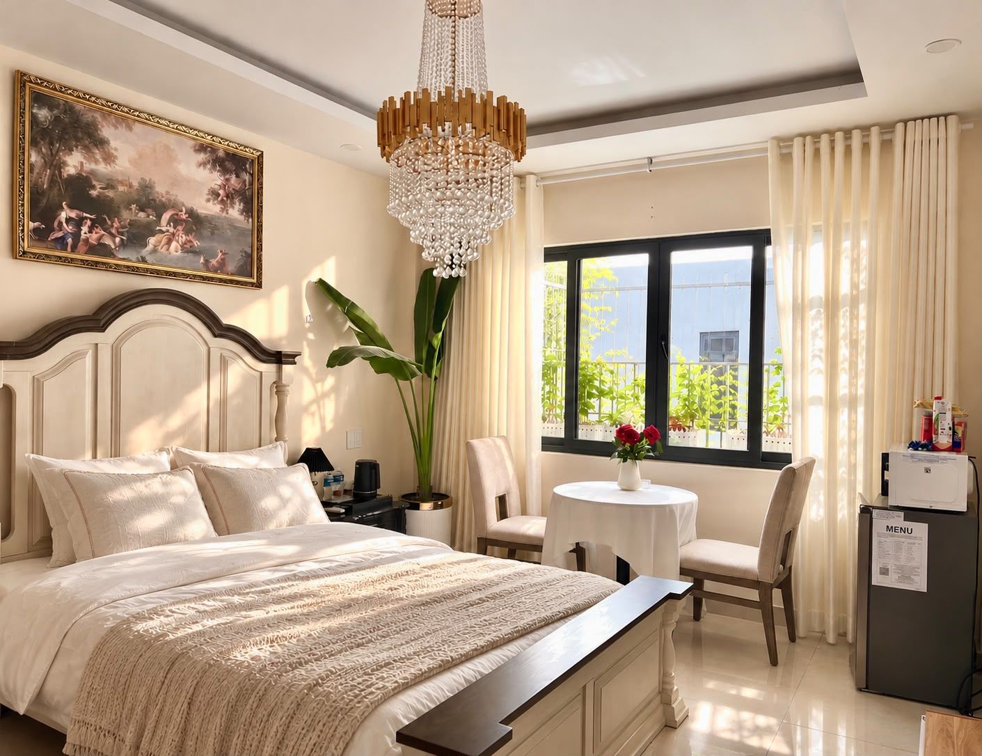 A hotel room with a bed, a desk, a chair, a dining table, a chandelier, a window with curtains, and a painting on the wall.