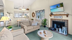 Un salon avec un canapé beige, une table basse, une cheminée et un téléviseur diffusant une scène de plage.
