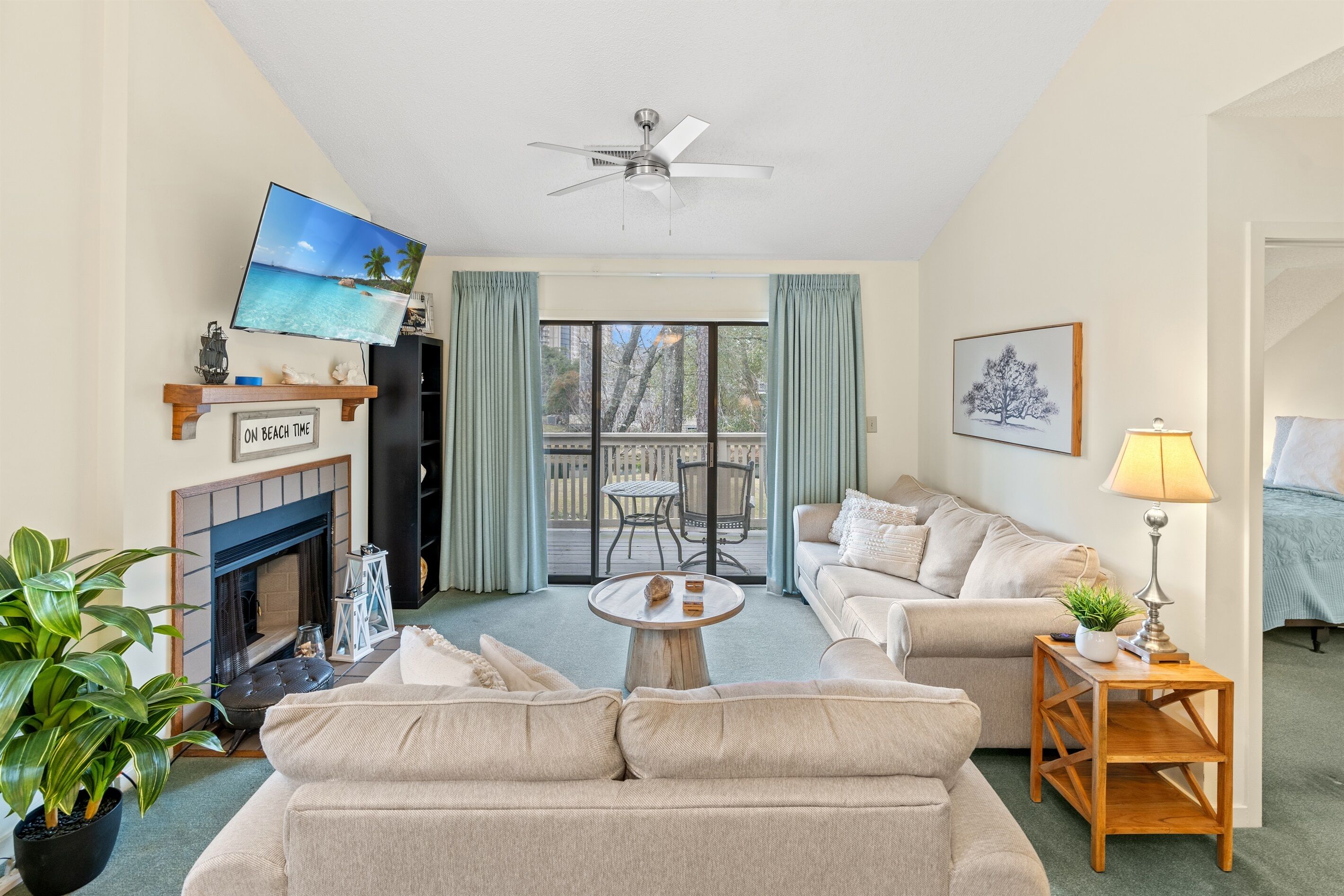 Un salon avec un canapé beige, une table basse, un téléviseur fixé au mur, un ventilateur de plafond, une cheminée et un balcon avec vue.