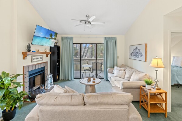 Un salon avec un canapé beige, une table basse, un téléviseur fixé au mur, un ventilateur de plafond, une cheminée et un balcon avec vue.