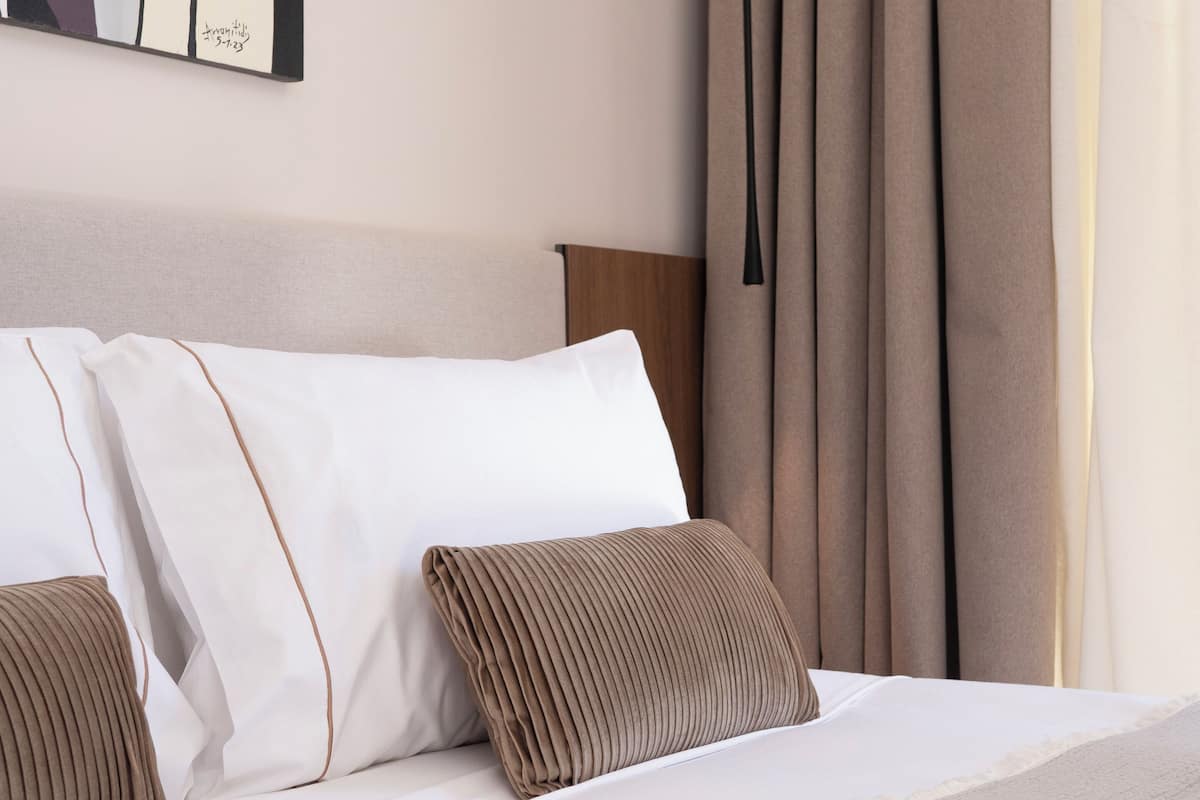 A neatly made bed with white linens and two brown pillows, a wooden headboard, and a painting on the wall.