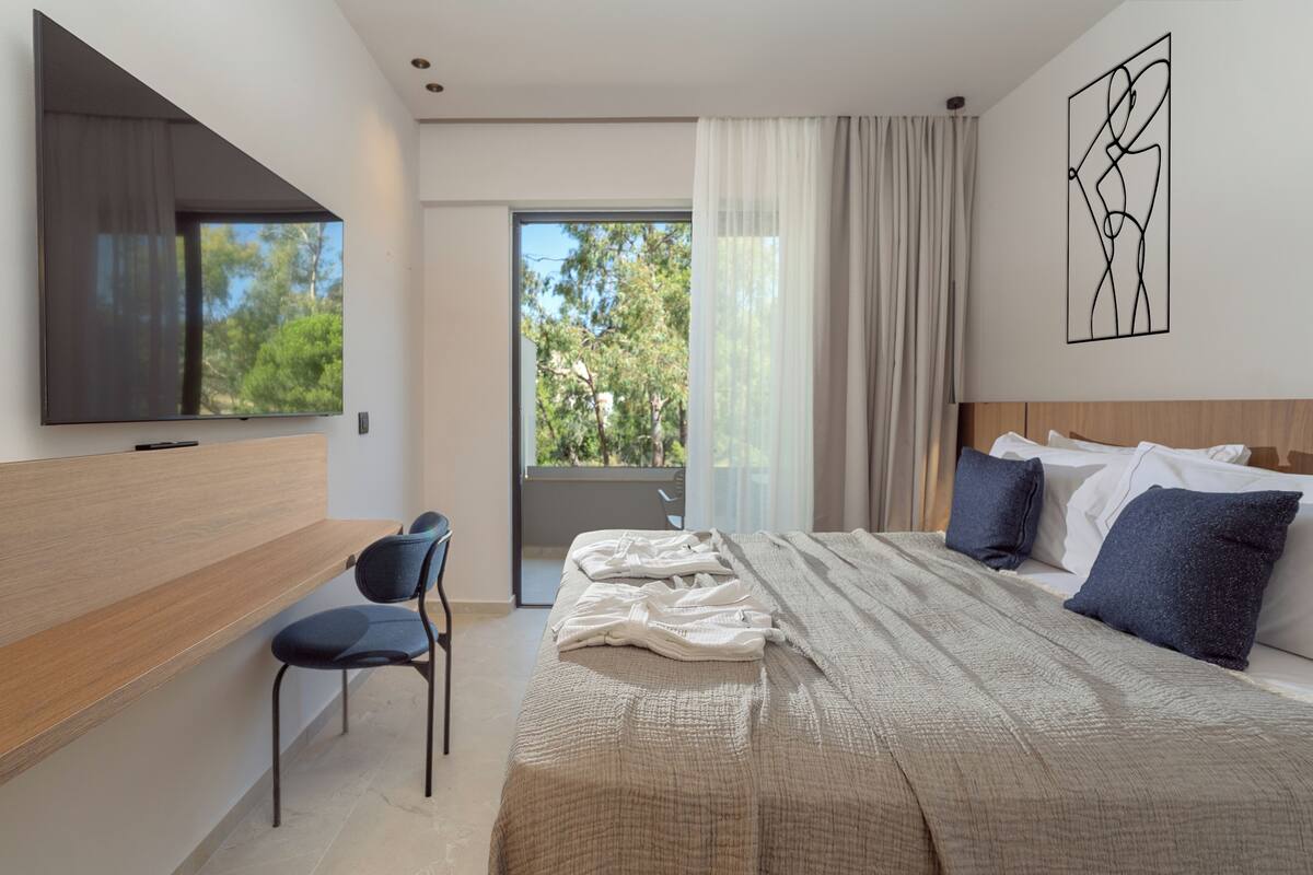 A modern hotel room with a large bed, a desk with a chair, a TV, and a balcony with a view of trees.