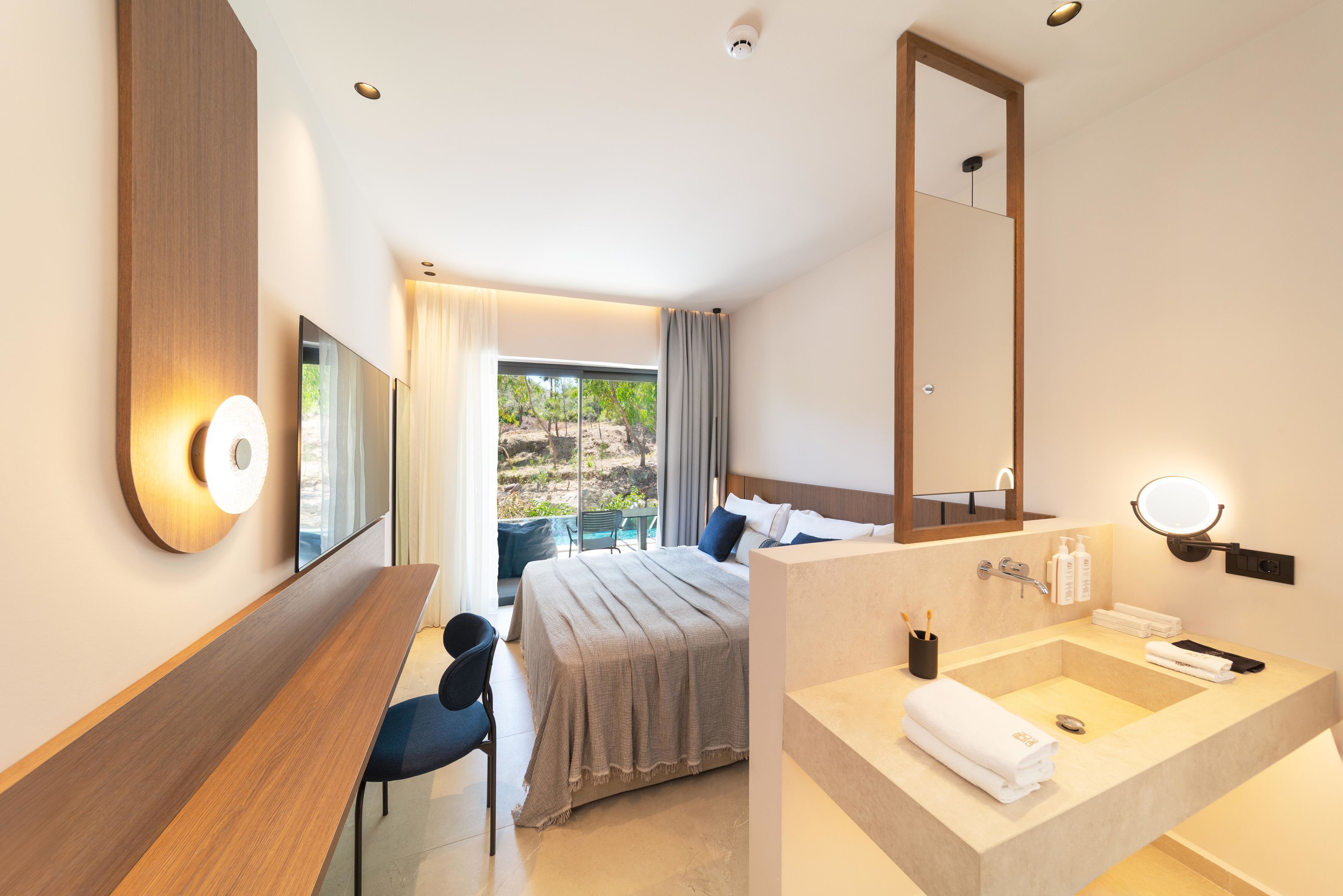 A modern hotel room with a bed, a desk, a large mirror, and a bathroom with a sink and shower.