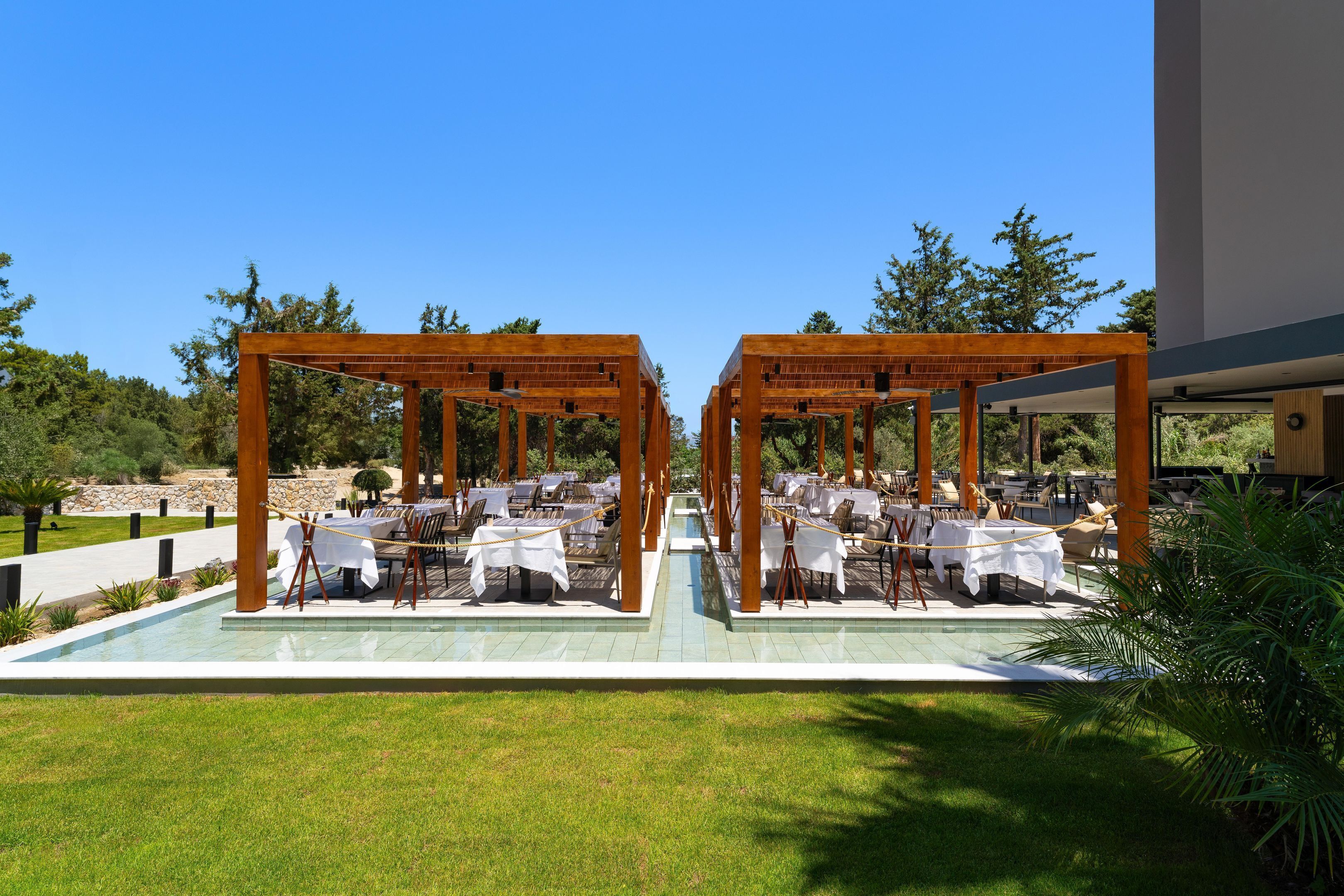 An outdoor dining area with tables and chairs under wooden pergolas, surrounded by greenery and a pool.