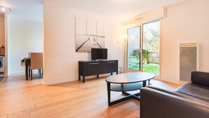 A modern living room with hardwood flooring, a black sofa, a round coffee table, a black TV stand, and a sliding glass door leading to a backyard.