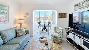 A living room with a sofa, coffee table, armchair, TV, and a dining area with chairs and a table. - Waterfront Condo 2BR/2BA, HEATED Pool, Short Walk to Beach, Dolphins & Manatees (Treasure Island)