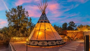 Un tipi con un diseño geométrico, rodeado por un sendero de piedra y una valla de madera, ubicado en un área boscosa.