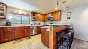 Une cuisine moderne avec des meubles en bois, des appareils électroménagers en acier inoxydable, un grand îlot central doté de tabourets et une fenêtre offrant une vue sur les arbres.