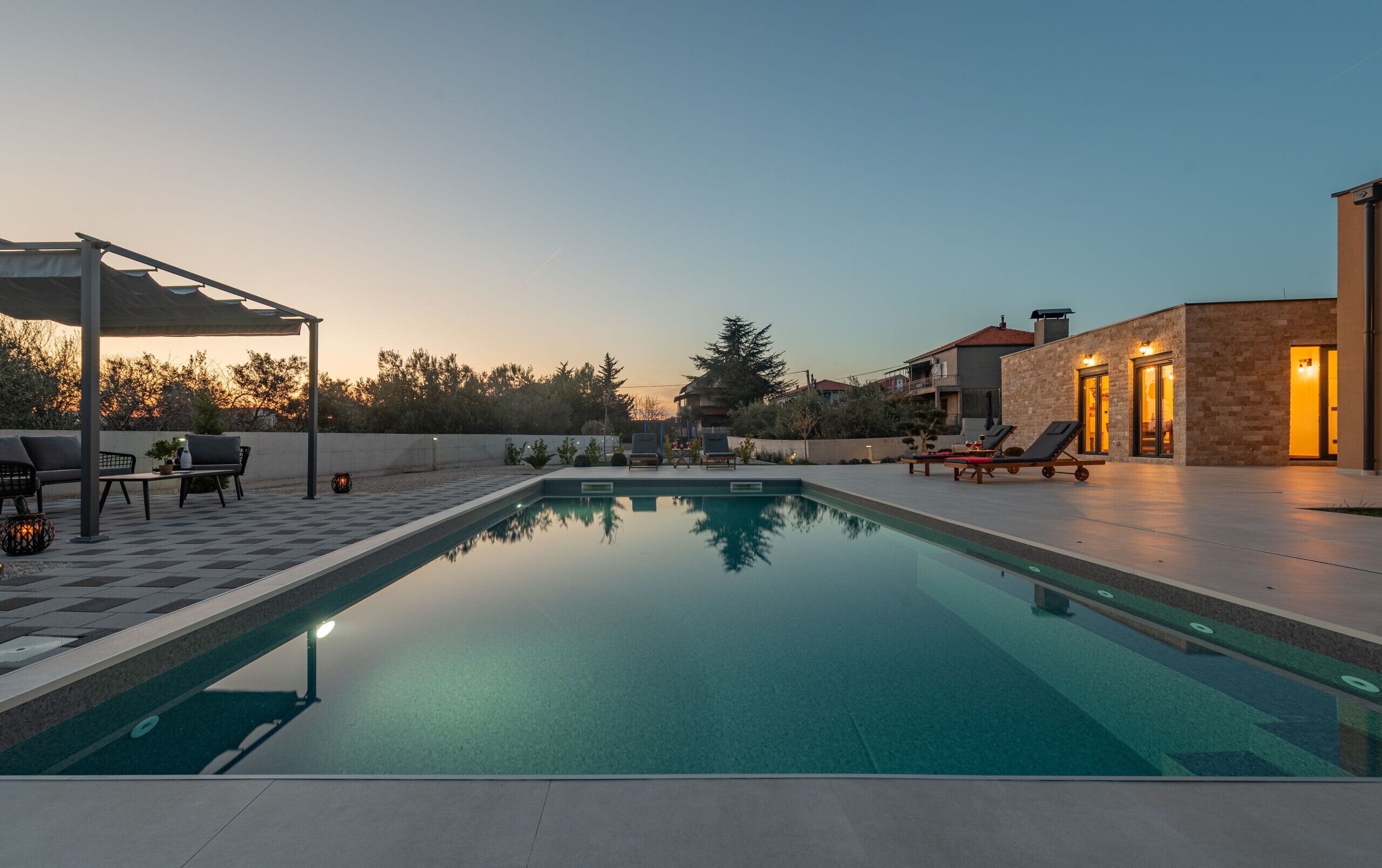 A swimming pool with lounge chairs, a covered seating area, and a building with a lit entrance.