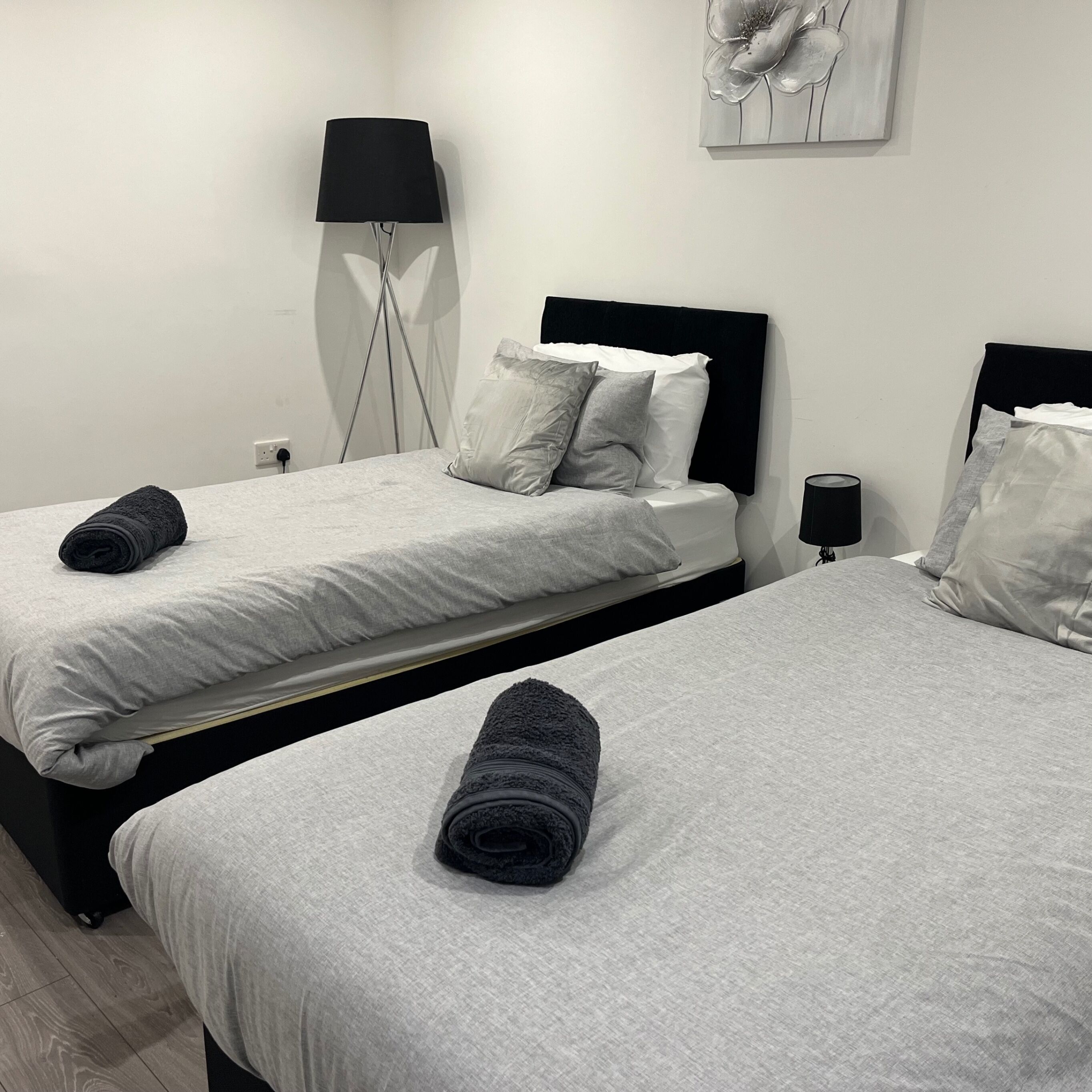 Habitación de hotel con dos camas, cada una con cabecera negra, ropa de cama blanca y una toalla enrollada sobre la cama. Hay una lámpara de pie y una obra de arte colgada en la pared sobre las camas.