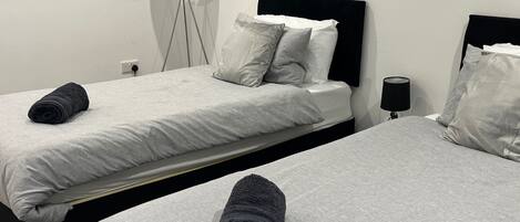 Habitación de hotel con dos camas, cada una con cabecera negra, ropa de cama blanca y una toalla enrollada sobre la cama. Hay una lámpara de pie y una obra de arte colgada en la pared sobre las camas.