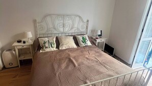A neatly made bed with a white metal frame, a brown quilt, and decorative pillows. There are two bedside tables with lamps and a small shelf. The room has a large window on the right side. - Bocca di rosa in centro (La Spezia)