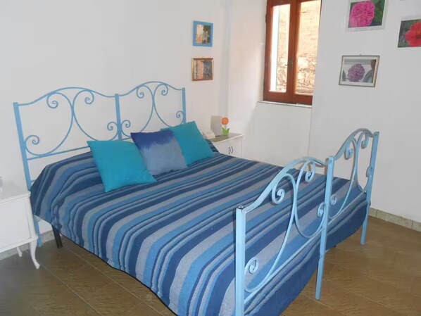 A bed with a blue metal frame and striped bedding, a bedside table with a lamp, and a window with a view of the outside. - Scirocco a Riomaggiore (Riomaggiore)