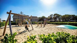 Ein Strandbereich mit Sand, einer Holz-Schaukel und einer Rutsche, umgeben von Grünpflanzen und einem Gebäude mit geneigtem Dach.