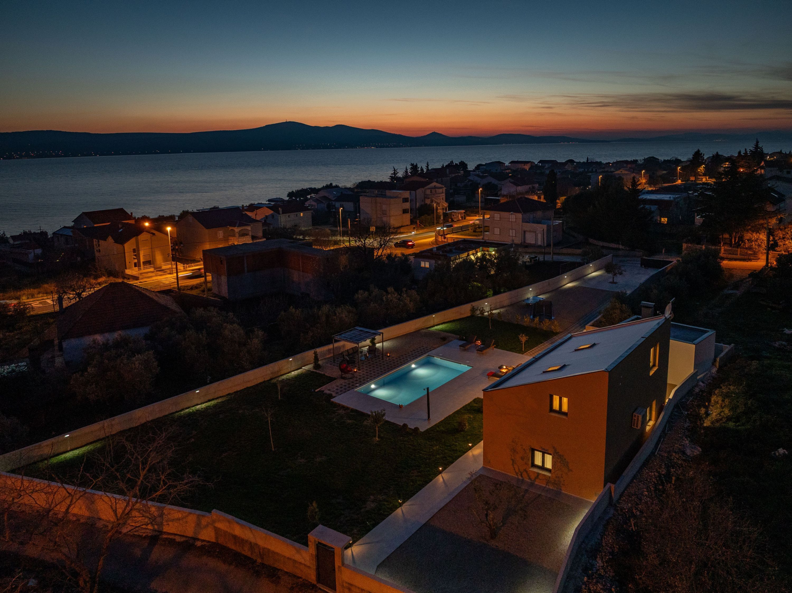 A residential area with a swimming pool, houses, and a view of the sea at dusk.