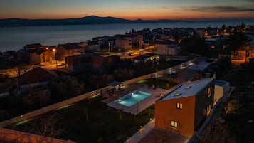 A residential area with a swimming pool, houses, and a view of the sea at dusk.