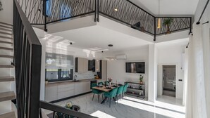 A spacious living area with a high ceiling, a dining table with chairs, a kitchen area, and a staircase with a railing.