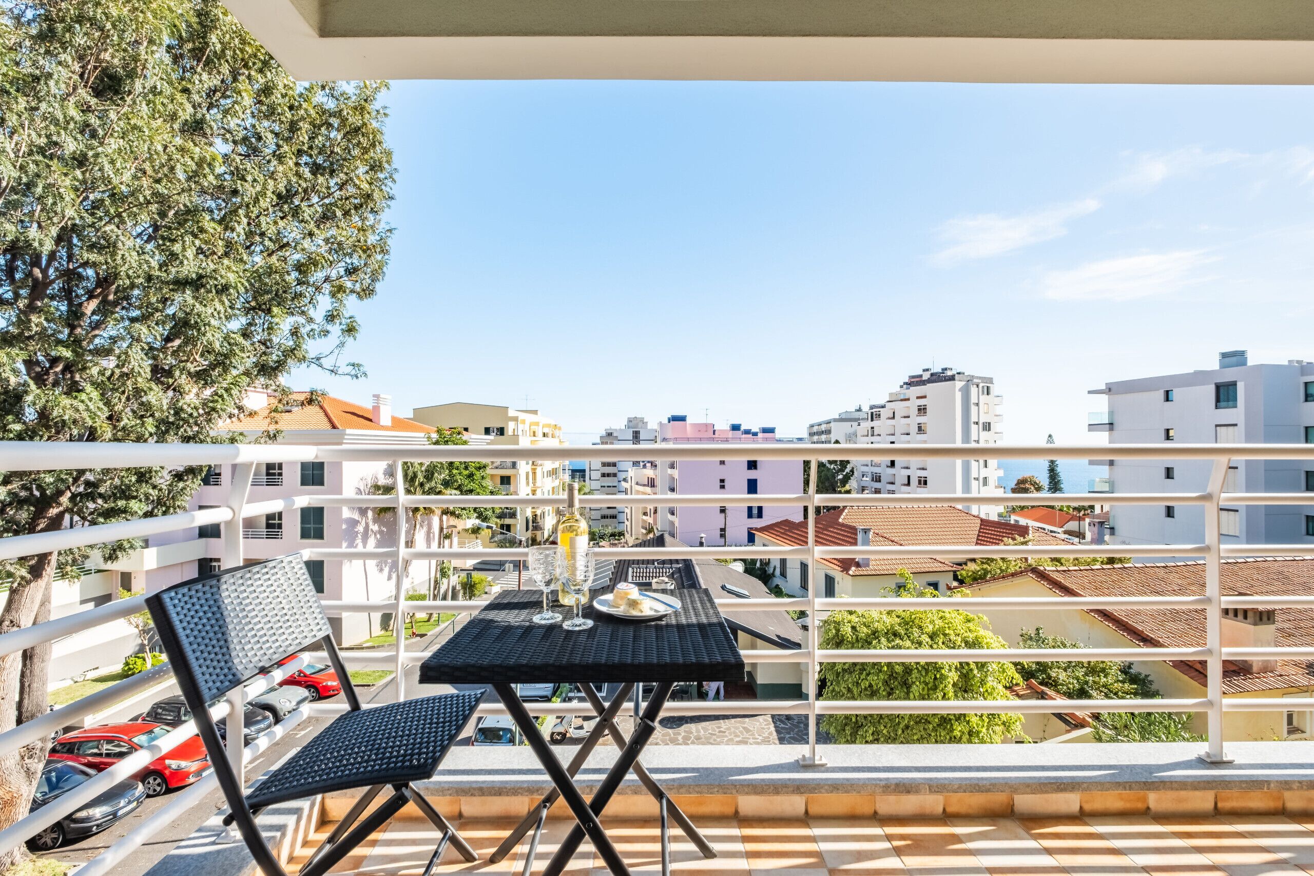 A balcony with a table and chairs, overlooking a residential area with multiple buildings and parked cars.