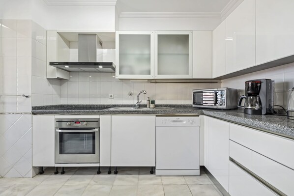 A modern kitchen with white cabinetry, a stainless steel oven, a dishwasher, a coffee maker, and a toaster. - Serenity Solace by Homie (Funchal)
