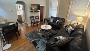 Una sala de estar con un sofá de cuero negro, una mesa de centro y un televisor en la pared. Hay un comedor con una mesa y sillas.