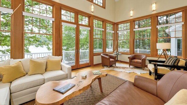 Una habitación amplia con ventanas grandes, un sofá blanco, un sofá de cuero marrón y una mesa de centro de madera.
