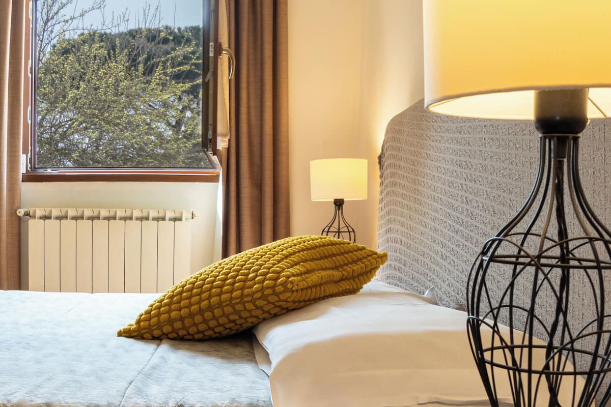 Un dormitorio con cama, lámpara de noche, ventana con cortinas y radiador.