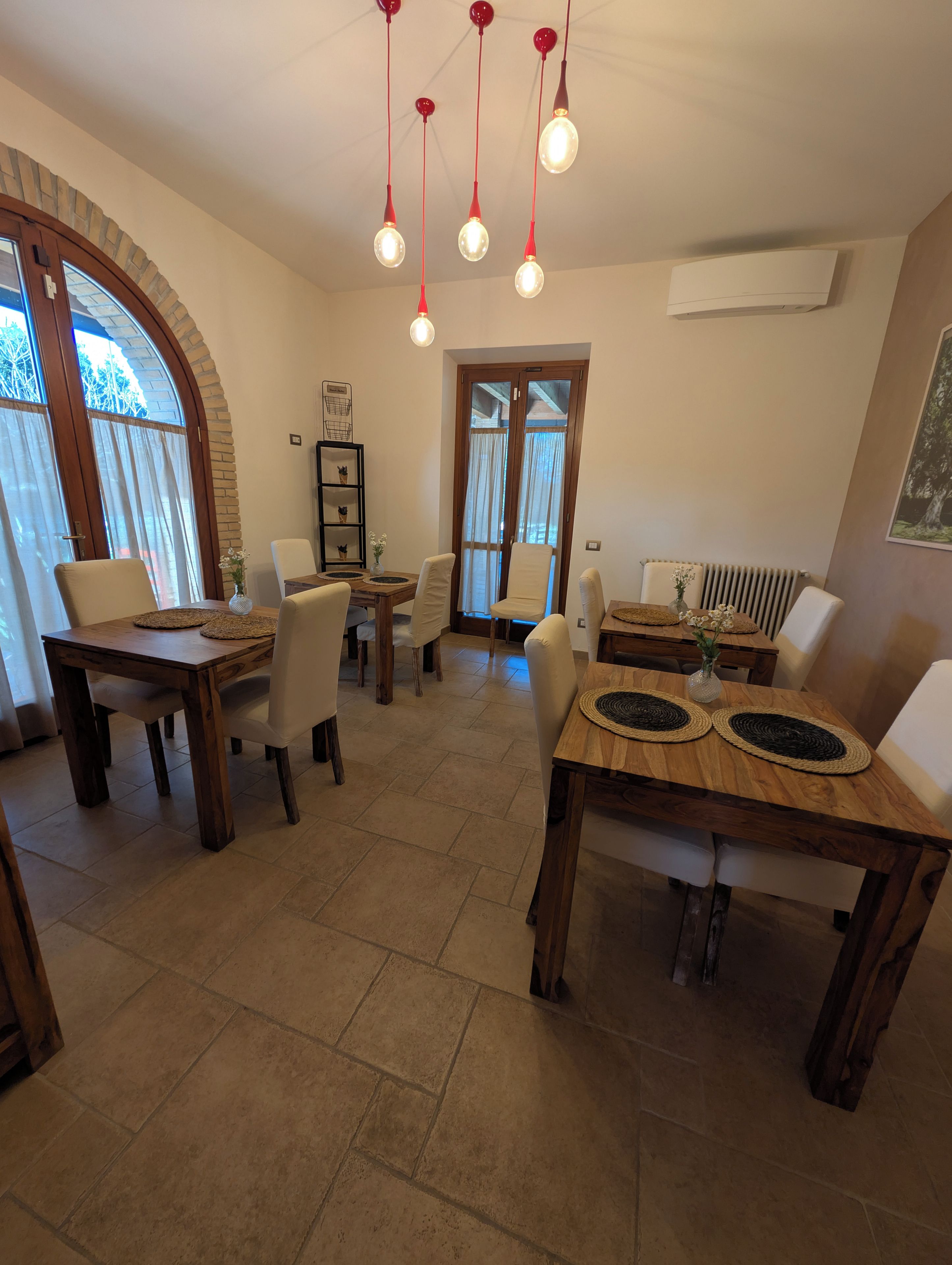 A dining area with wooden tables and chairs, a chandelier, and a view through a large arched window.