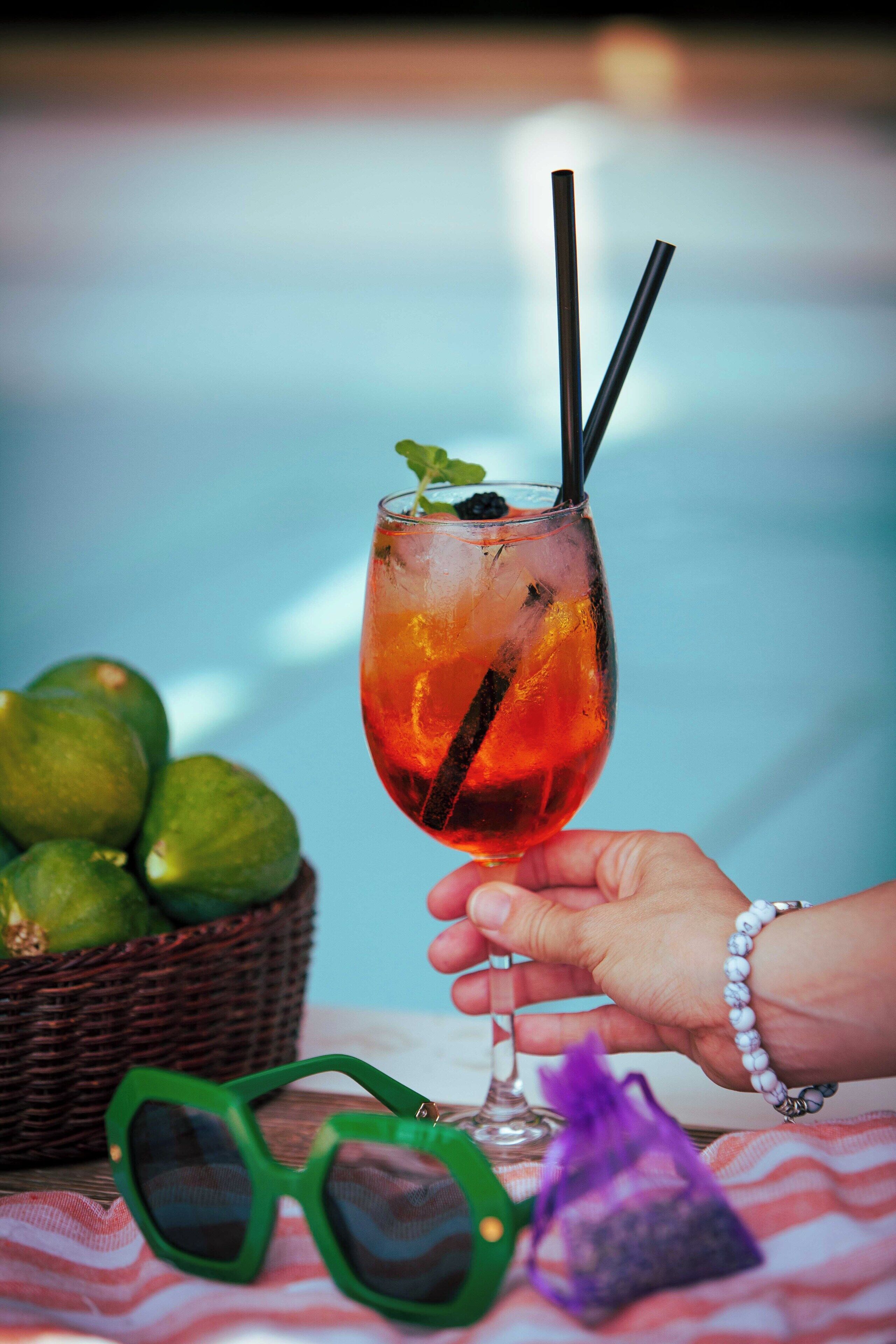 A hand holding a cocktail glass with a straw, a basket of limes, and green sunglasses on a table.
