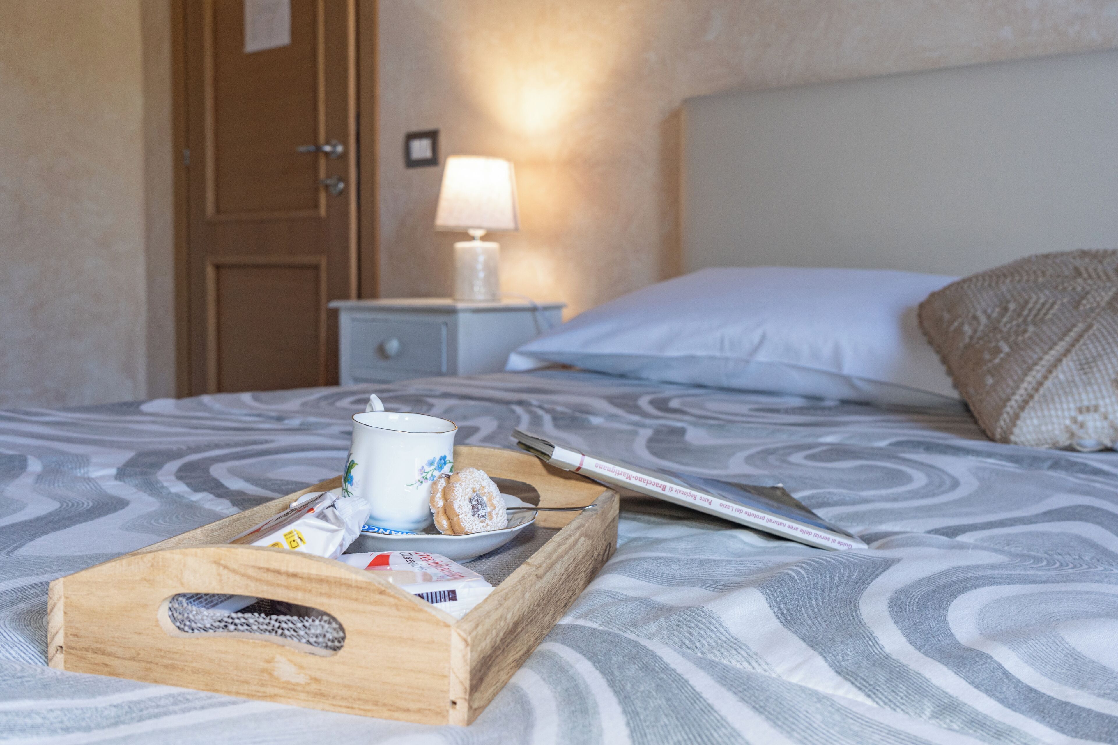 Una cama bien tendida con una bandeja de alimentos para el desayuno, una lámpara de noche y una mesita de madera.