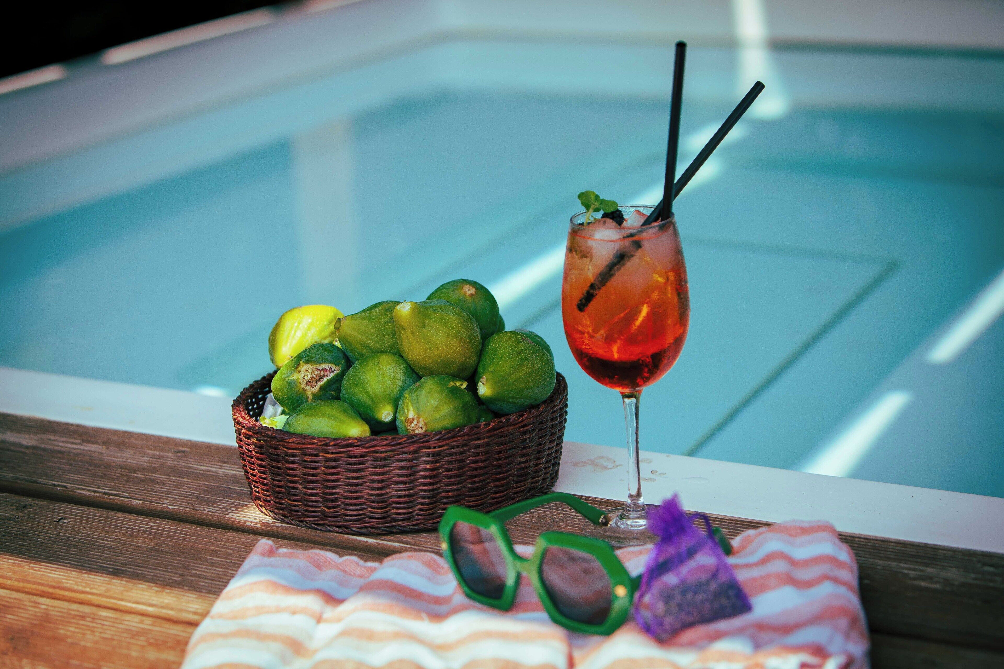 A poolside setting with a wicker basket of limes, a glass of red drink with straws, and a pair of sunglasses on a striped towel.