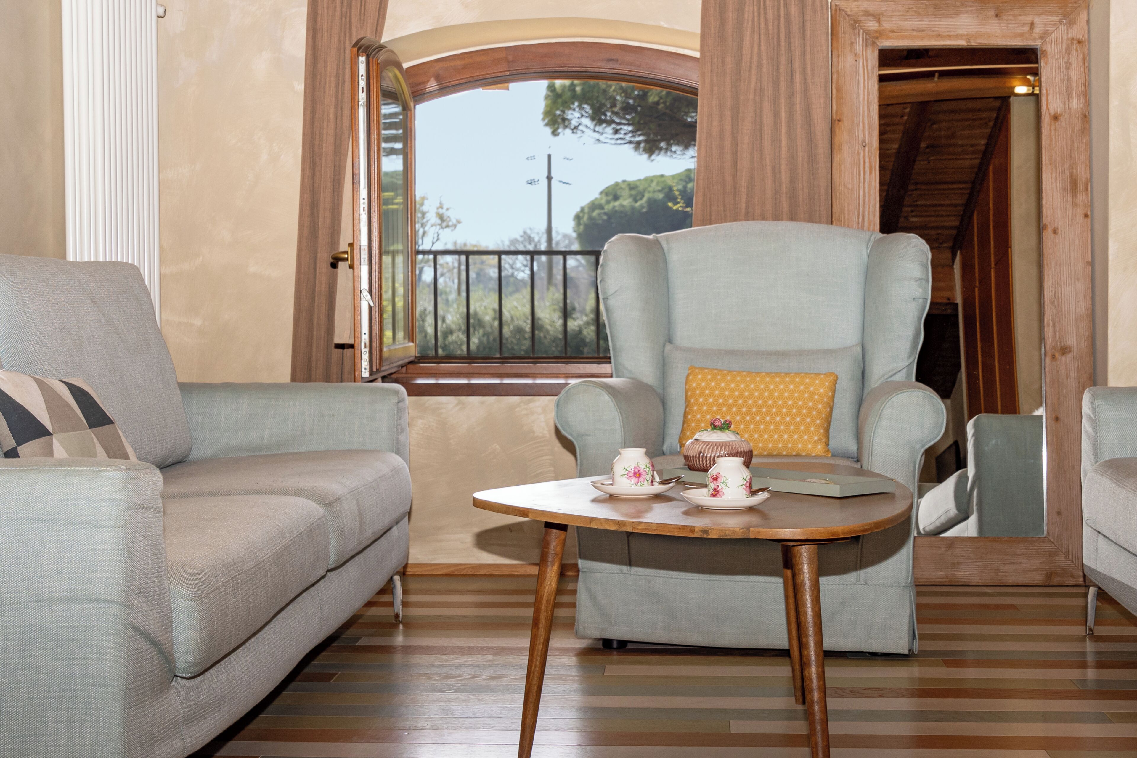 Une pièce comprenant un canapé, un fauteuil, une table avec des tasses à thé et une fenêtre donnant sur des arbres.