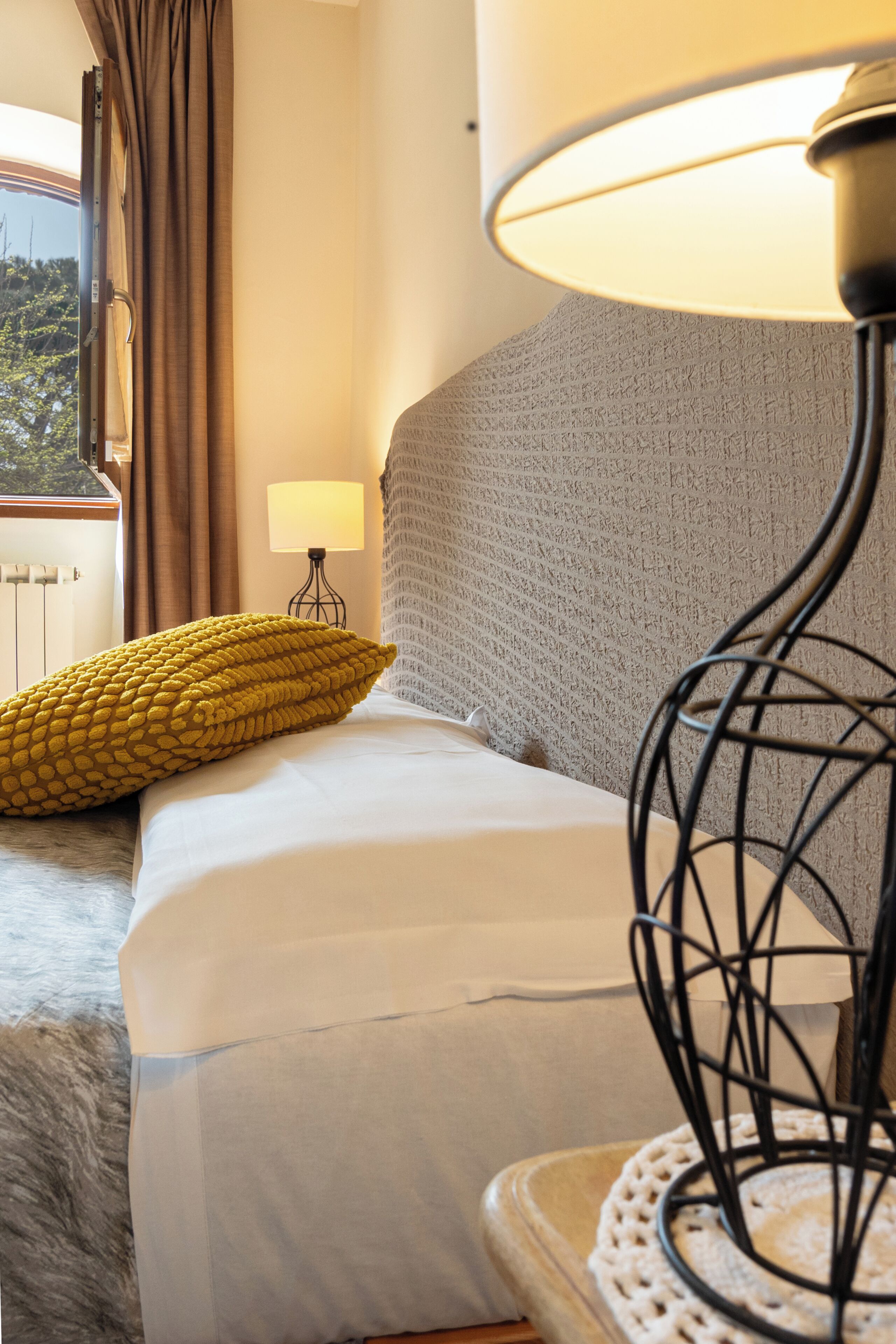 A hotel room with a bed, a lamp, a window with curtains, and a view of the outdoors.