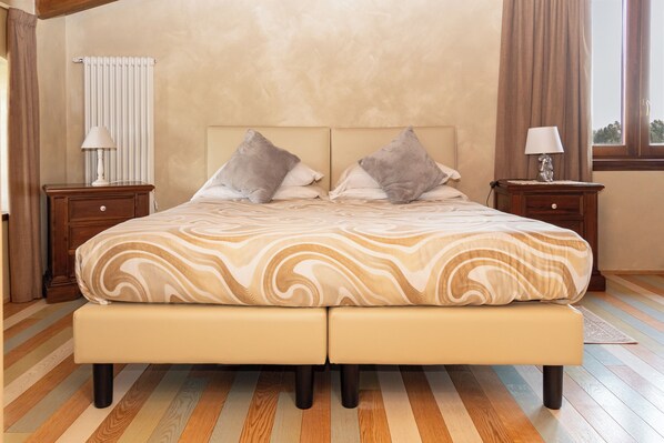 Una cama doble con una colcha de estampado marmolizado, dos mesitas de noche con lámparas, un radiador y una ventana con cortinas.