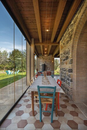 Un área de comedor al aire libre techada con un muro de piedra, vigas de madera y vistas al jardín.