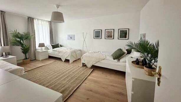 A modern, minimalist bedroom with a large bed, bedside tables, a standing lamp, and a potted plant.
